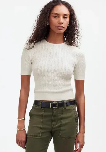 A woman with curly hair wearing a fitted short-sleeve cream knit top and olive green pants with a black belt stands confidently against a plain white background.