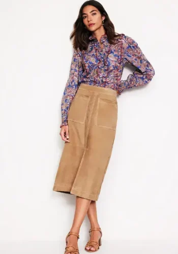 A woman wearing a patterned long-sleeve blouse paired with a tan midi skirt and brown heels poses stylishly against a white background.