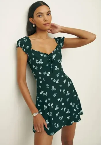 A woman wearing a green floral-print mini dress with puff sleeves poses with one hand behind her neck, exuding elegance and confidence against a light-colored background.