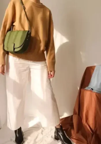 A person wearing a cozy tan sweater, wide-leg white pants, black ankle boots, and carrying a green crossbody bag poses stylishly in a softly lit room.