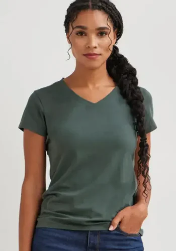 A woman wearing a green v-neck t-shirt and dark blue jeans stands confidently with a calm expression against a light-colored background.