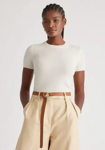 A woman wearing a white t-shirt, beige wide-leg pants with a brown belt, and brown shoes poses confidently with her hands in her pockets against a light-colored background.