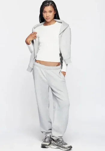 A woman wearing a white long-sleeve top, light gray sweatpants, and a matching gray hoodie draped over her shoulders poses confidently against a plain white background.