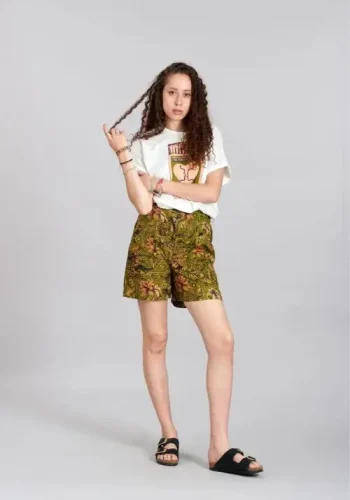 A woman wearing a graphic t-shirt and patterned green shorts poses playfully while holding a braid in her hair, standing against a light gray background. She completes the casual look with black slide sandals.