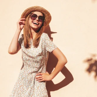 "A woman smiling and wearing a floral dress, wide-brimmed straw hat, and oversized sunglasses, showcasing elements of a summer capsule wardrobe."