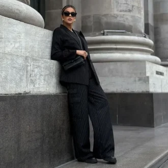 Fashionable woman in a pinstripe suit and black pants outfits leaning against a stone wall.