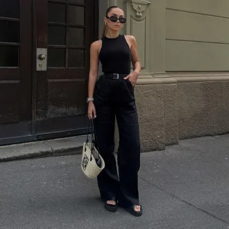 A stylish woman wearing women's black pants, paired with a sleeveless black top, black sandals, and a chic belt, accessorized with sunglasses and a designer handbag, standing confidently in an urban setting.