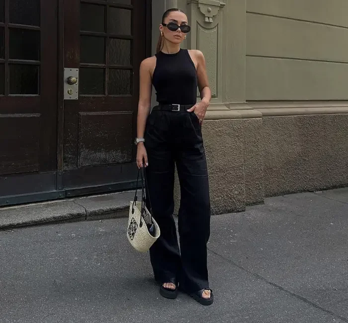 A stylish woman wearing women's black pants, paired with a sleeveless black top, black sandals, and a chic belt, accessorized with sunglasses and a designer handbag, standing confidently in an urban setting.