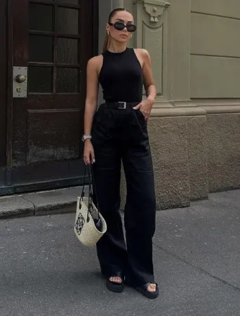 A stylish woman wearing women's black pants, paired with a sleeveless black top, black sandals, and a chic belt, accessorized with sunglasses and a designer handbag, standing confidently in an urban setting.