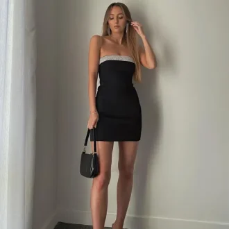 Young woman wearing a sleek, strapless little black dress paired with black heels and a small handbag, standing against a minimalist indoor background. The little black dress is a timeless and versatile piece that every woman should have in her wardrobe.