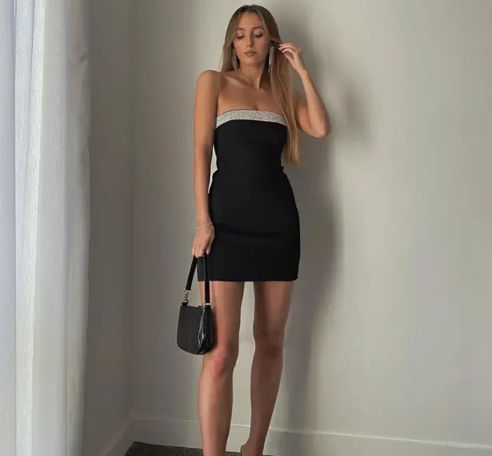Young woman wearing a sleek, strapless little black dress paired with black heels and a small handbag, standing against a minimalist indoor background. The little black dress is a timeless and versatile piece that every woman should have in her wardrobe.