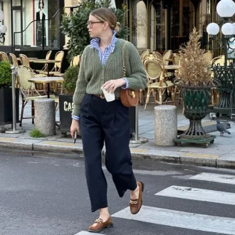 Woman wearing a casual chic dress code outfit consisting of a green knit sweater over a striped blue button-up shirt, high-waisted navy trousers, brown loafers with gold chain detail, and a brown crossbody bag, walking on a city street.