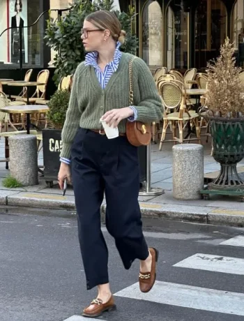 Woman wearing a casual chic dress code outfit consisting of a green knit sweater over a striped blue button-up shirt, high-waisted navy trousers, brown loafers with gold chain detail, and a brown crossbody bag, walking on a city street.