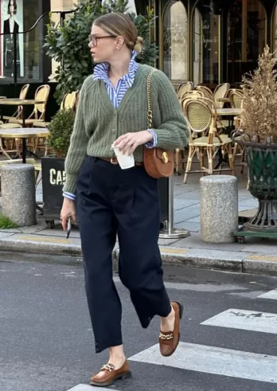 Woman wearing a casual chic dress code outfit consisting of a green knit sweater over a striped blue button-up shirt, high-waisted navy trousers, brown loafers with gold chain detail, and a brown crossbody bag, walking on a city street.