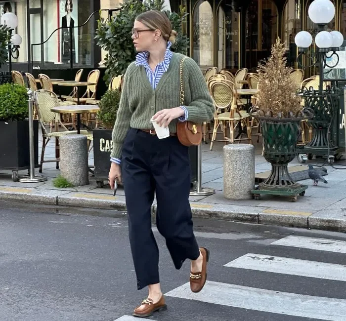 Woman wearing a casual chic dress code outfit consisting of a green knit sweater over a striped blue button-up shirt, high-waisted navy trousers, brown loafers with gold chain detail, and a brown crossbody bag, walking on a city street.