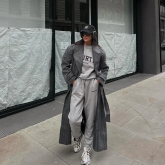 Girl in athleisure fashion, sporting a casual yet chic ensemble with baggy sweats, a gray shirt, a dark gray trench coat, and white sneakers, complemented by sunglasses and a hat.