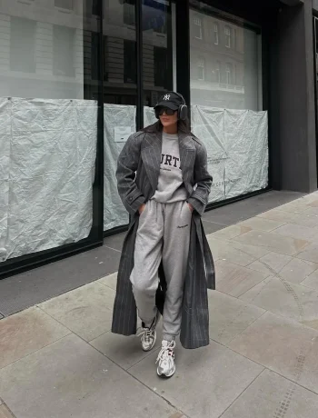 Girl in athleisure fashion, sporting a casual yet chic ensemble with baggy sweats, a gray shirt, a dark gray trench coat, and white sneakers, complemented by sunglasses and a hat.