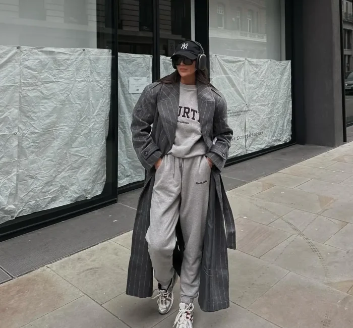 Girl in athleisure fashion, sporting a casual yet chic ensemble with baggy sweats, a gray shirt, a dark gray trench coat, and white sneakers, complemented by sunglasses and a hat.