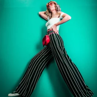 A young woman, showcasing seasonal styles, stands gracefully against a backdrop of lush, vibrant greenery. Her outfit consists of loose-fitting pants adorned with chic stripes, perfectly complemented by a cropped white shirt with a trendy short cut.