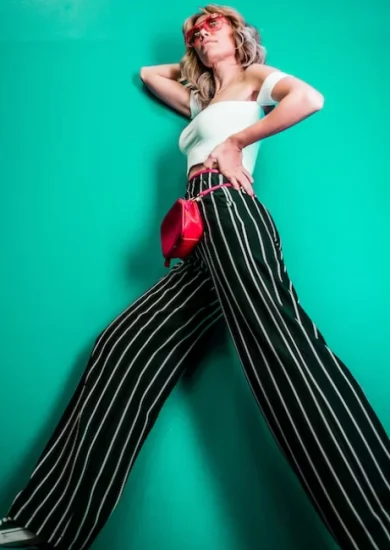 A young woman, showcasing seasonal styles, stands gracefully against a backdrop of lush, vibrant greenery. Her outfit consists of loose-fitting pants adorned with chic stripes, perfectly complemented by a cropped white shirt with a trendy short cut.