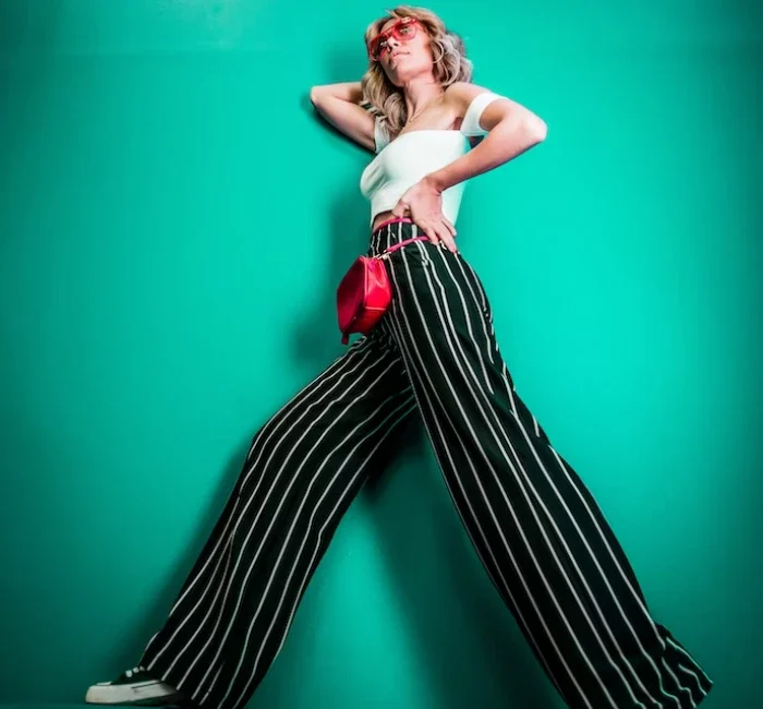 A young woman, showcasing seasonal styles, stands gracefully against a backdrop of lush, vibrant greenery. Her outfit consists of loose-fitting pants adorned with chic stripes, perfectly complemented by a cropped white shirt with a trendy short cut.