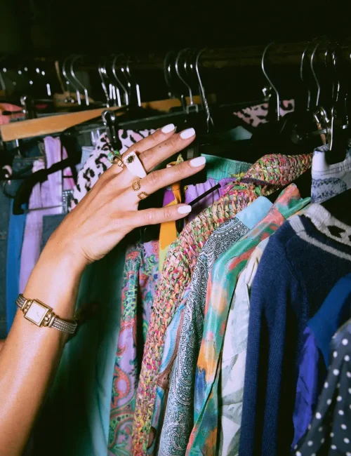 A close-up of a hand with rings and a watch, browsing through colorful, patterned clothing on a rack, showcasing a vibrant and eclectic fashion selection.