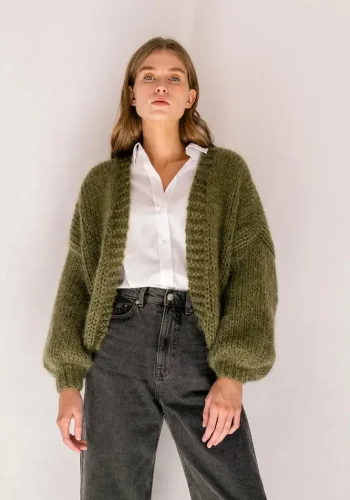 A woman wearing an olive green chunky knit cardigan over a white button-up shirt, paired with high-waisted black jeans, stands confidently against a neutral background.