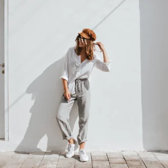 Stylish girl in gray pants and white cotton blouse posing near white wall, showcasing a Seasonal Minimalist Wardrobe Transition look. Woman in cap and glasses.