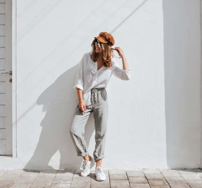 Stylish girl in gray pants and white cotton blouse posing near white wall, showcasing a Seasonal Minimalist Wardrobe Transition look. Woman in cap and glasses.