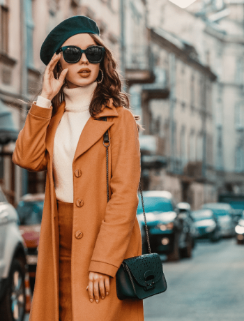 A stylish young woman showcasing her personal style in an outdoor fall portrait, wearing trendy sunglasses, a camel-colored coat, turtleneck, and a textured leather shoulder bag, strolling through the streets of a European city.