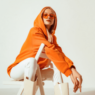 Stylish young blonde woman in a vibrant orange hoodie and colorful sunglasses, striking a pose against a white backdrop in a studio fashion portrait