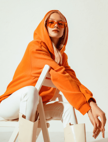 Stylish young blonde woman in a vibrant orange hoodie and colorful sunglasses, striking a pose against a white backdrop in a studio fashion portrait