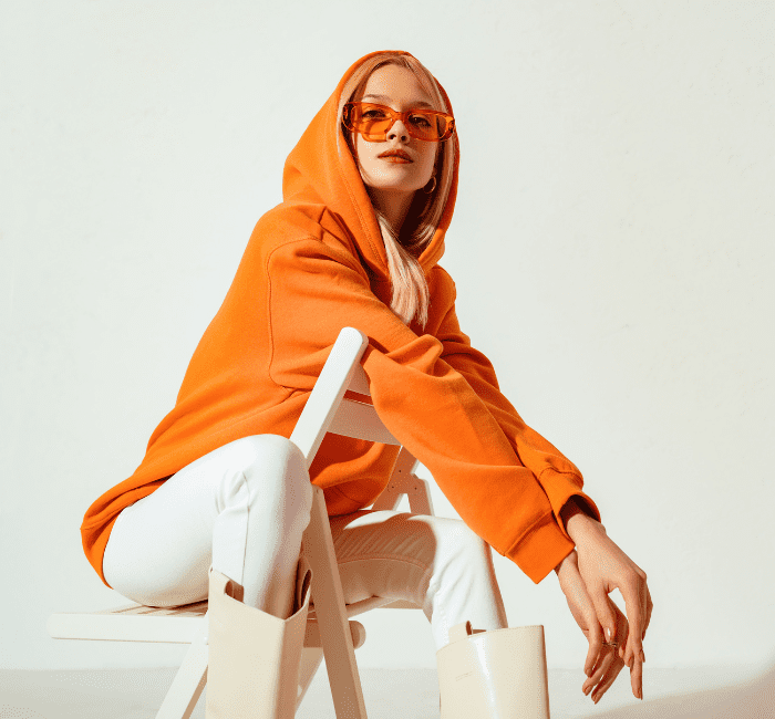 Stylish young blonde woman in a vibrant orange hoodie and colorful sunglasses, striking a pose against a white backdrop in a studio fashion portrait