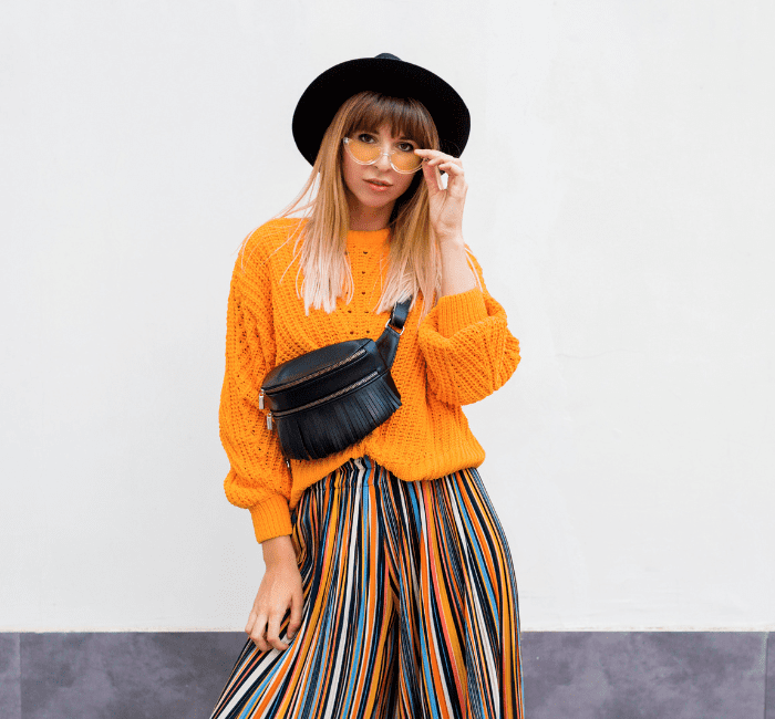 Cheerful brunette woman defying fashion rules, standing on white background in trendy orange sweater and multicolor striped culottes