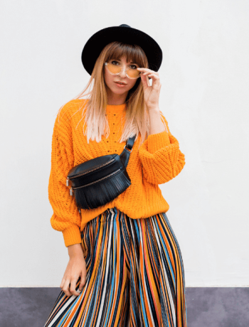 Cheerful brunette woman defying fashion rules, standing on white background in trendy orange sweater and multicolor striped culottes