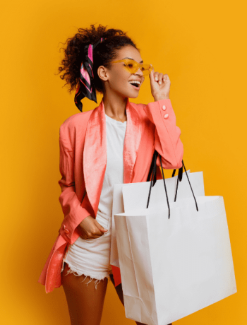 Studio shot of a beautiful black woman holding a white shopping bag, standing against a yellow background, embodying a trendy spring look and providing a visual shopping guide inspiration.