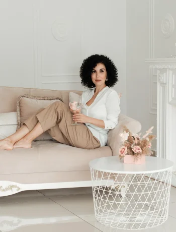 Woman with curly hair sits on a sofa with bare feet and holds a glass of water enjoying minimalist living