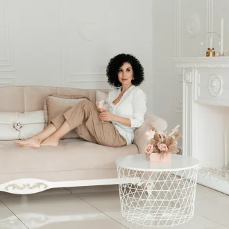 Woman with curly hair sits on a sofa with bare feet and holds a glass of water enjoying minimalist living